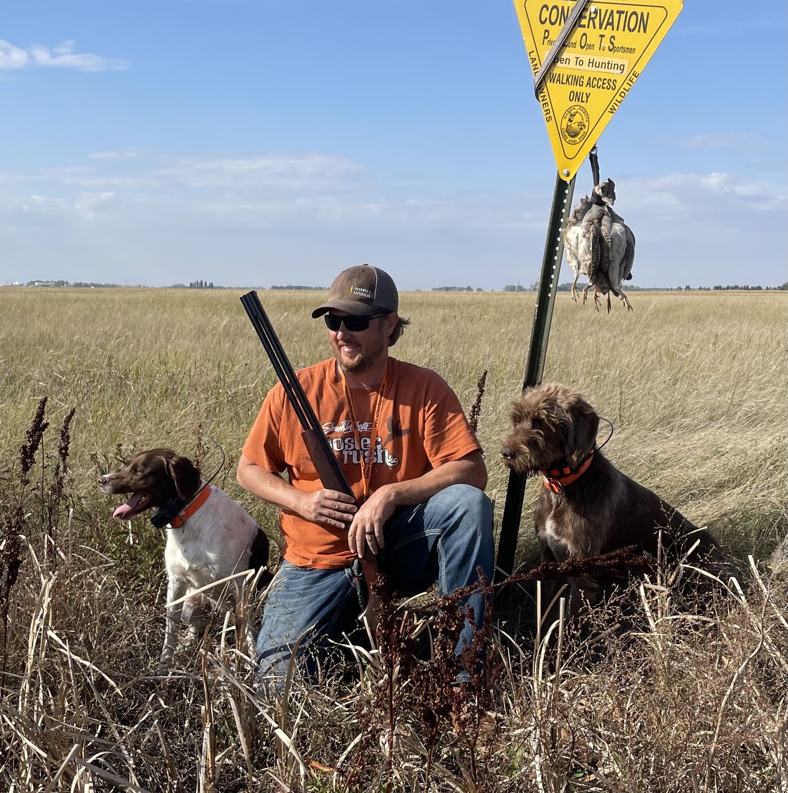 Dave with two dogs and a shotgun beside a walk-in conservation area sign, sharp-tailed grouse hanging from the sign